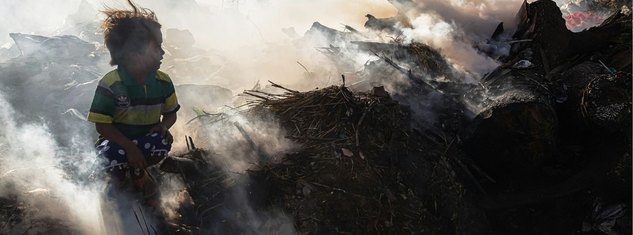 Photograph of a child sitting amid smouldering piles of waste at a landfill, surrounded by thick smoke and debris, with hazy light filtering through the air. It is depicting environmental pollution.