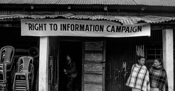 A black and white photograph that shows the entrance to a tin roofed structure with a banner on the top saying "Right to Information Campaign". Two women are standing close together to the right side of the image, outside the structure. Another man is standing in the doorway