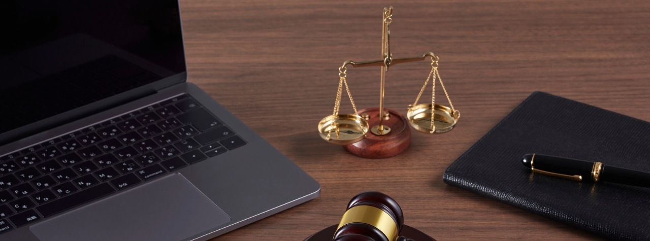 An open grey laptop, a gold-and-wood scales of justice, a closed black notebook with a fountain pen, and the top of a legal gavel arranged on a dark wooden desk.